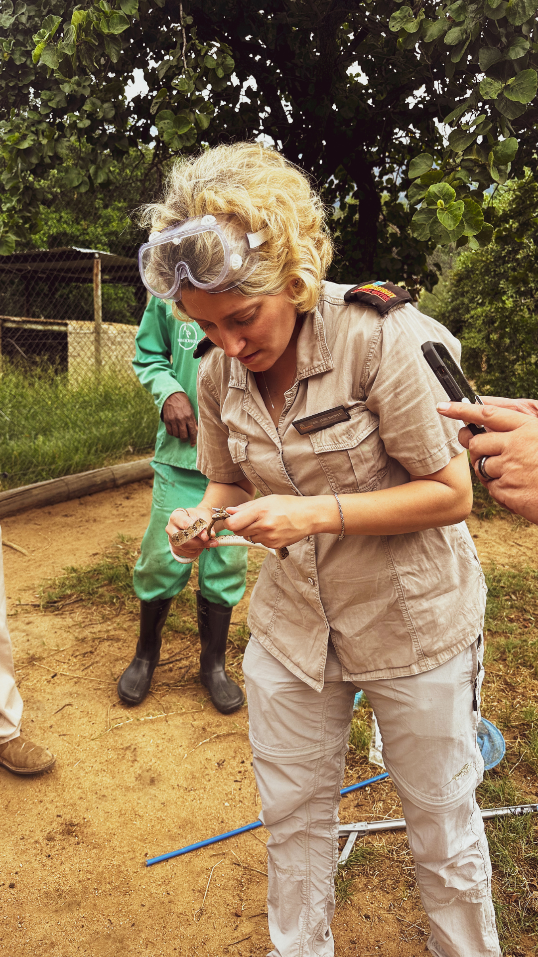 Woman in khaki uniform and safety goggles examining an object in a natural setting, with another person nearby.