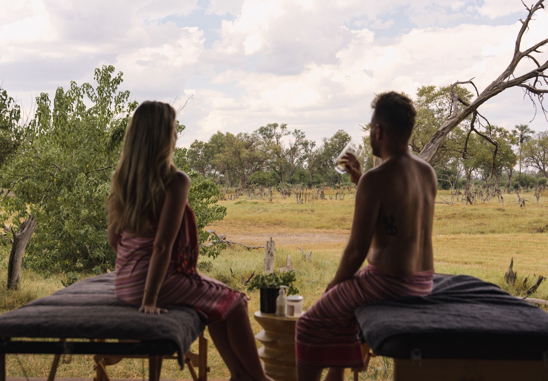Couple relaxing on massage tables outdoors, wrapped in towels, looking at the landscape.