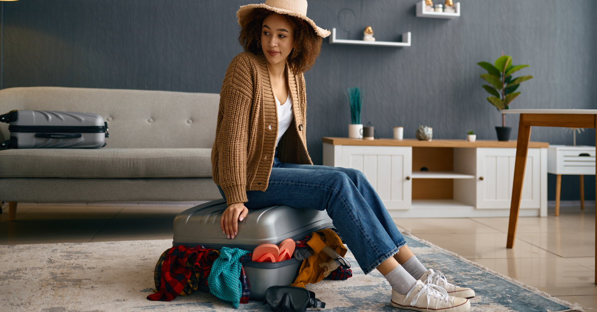 Woman struggling to close a suitcase, sitting on top in a living room.