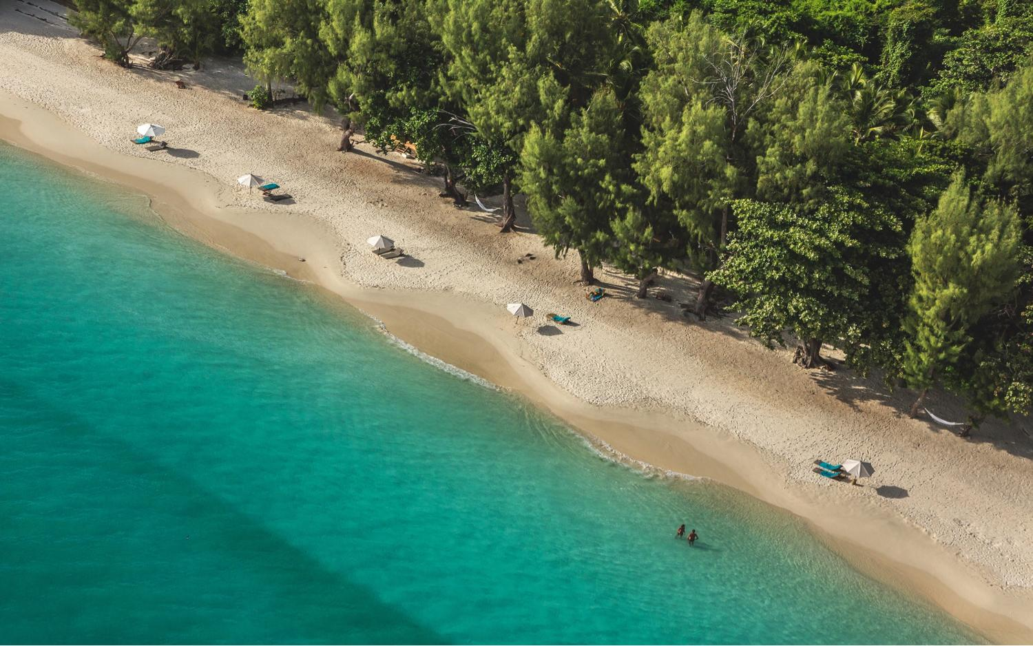 Sandy beach with turquoise water, trees, and a few people swimming.