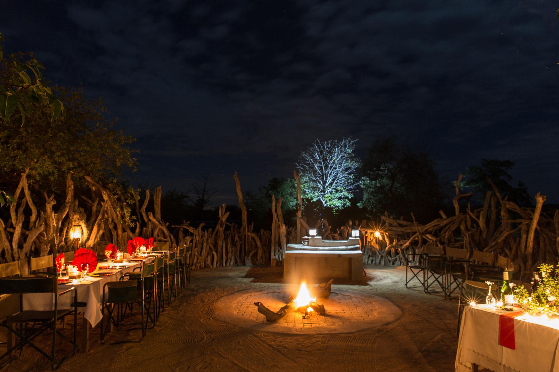 Nighttime outdoor dining setting with a bonfire, tables, chairs, and lanterns in a natural environment.