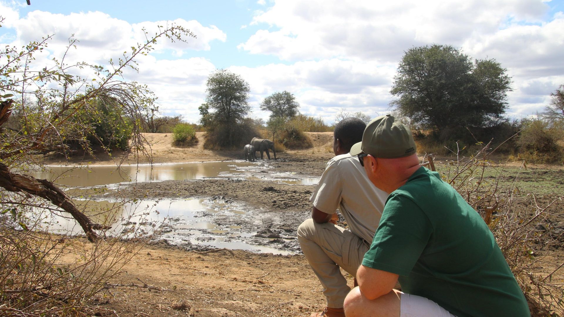 Two men watch an elephant at a watering hole in a savanna. The men are crouched down; the sky is partly cloudy.