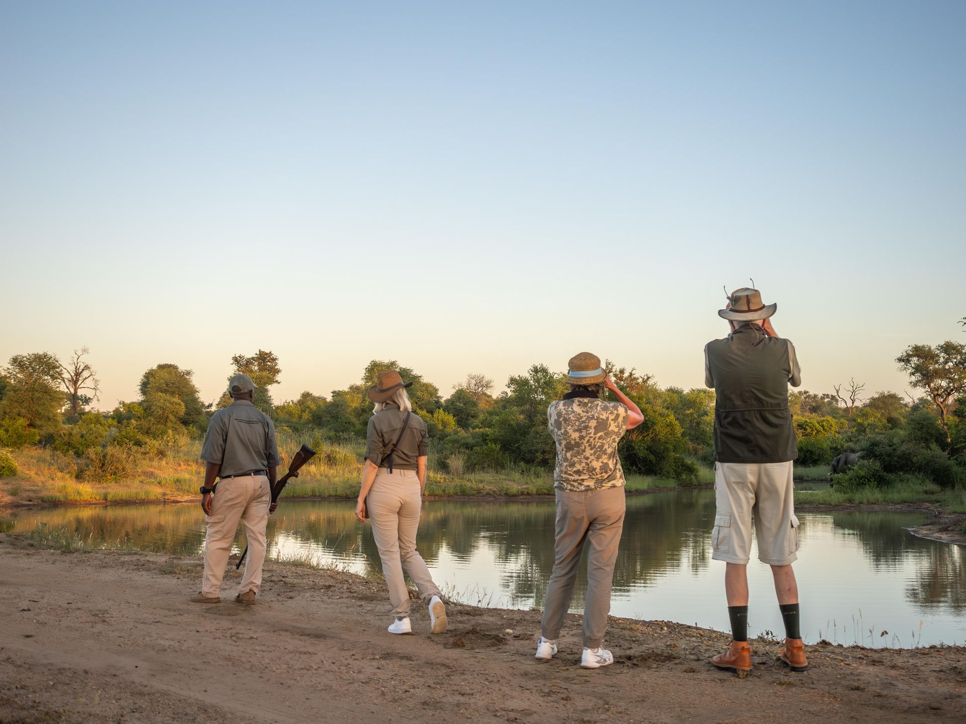 Four people in hats stand by a river at sunset, looking across the water and photographing wildlife.