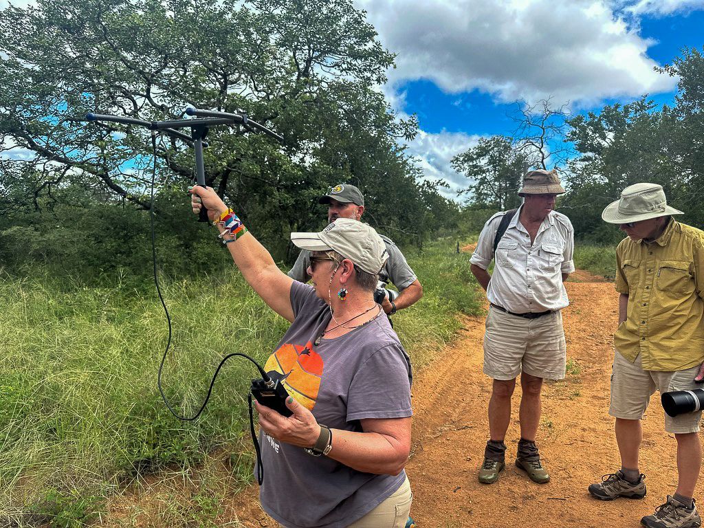 Woman operates a device, possibly for surveying, on a dirt path with three men observing in a natural setting.