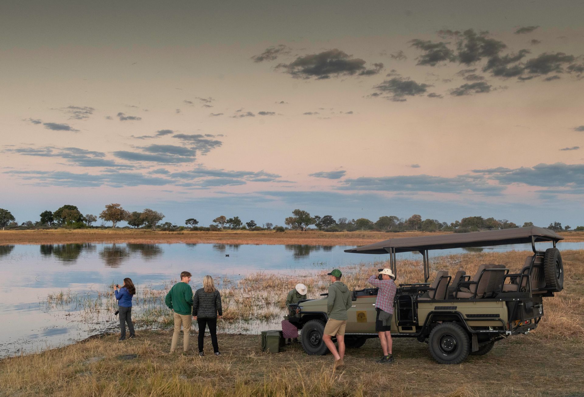 Safari vehicle with tourists near water at sunset.