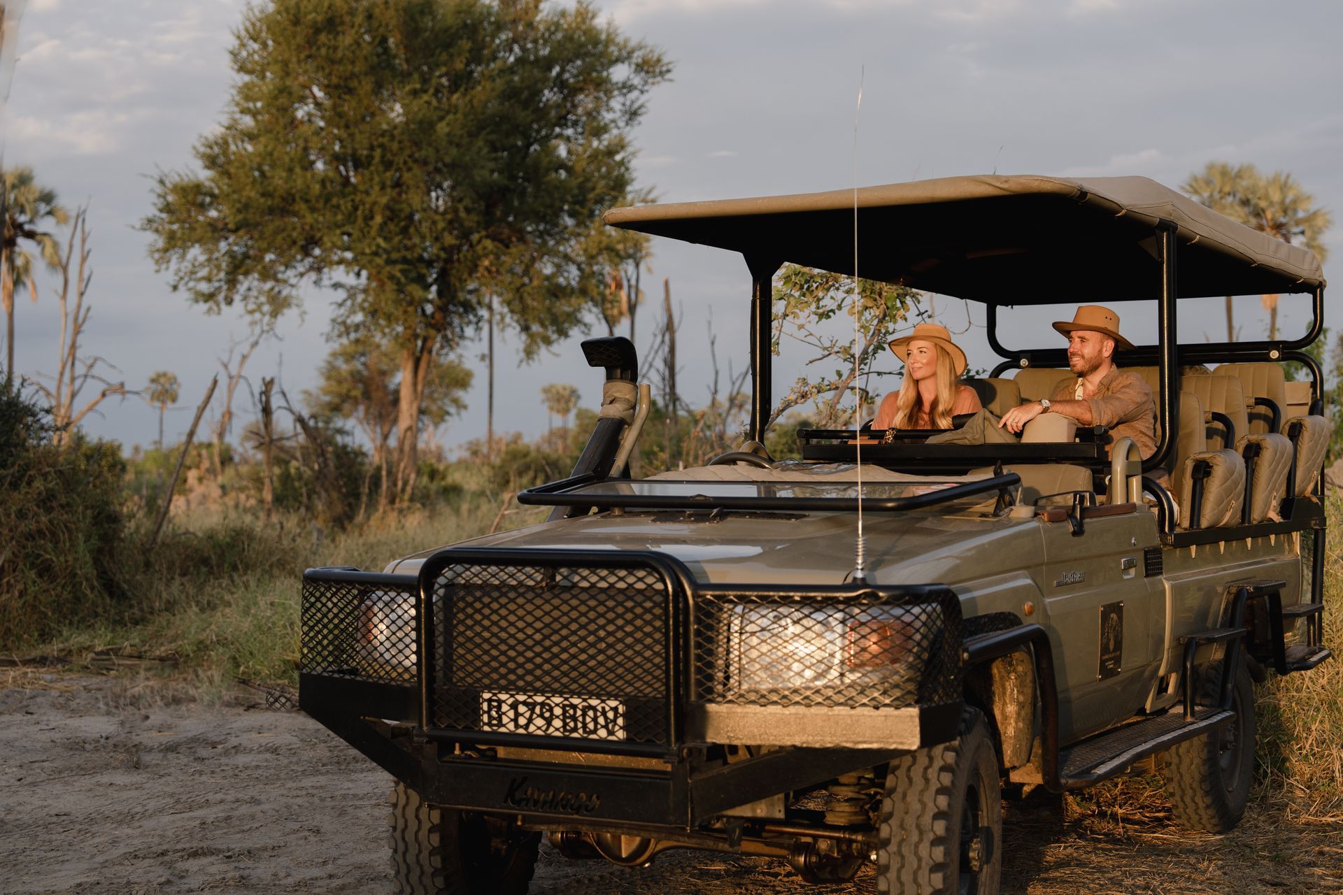 Safari vehicle with two people, driving through a savanna. Green and beige tones.