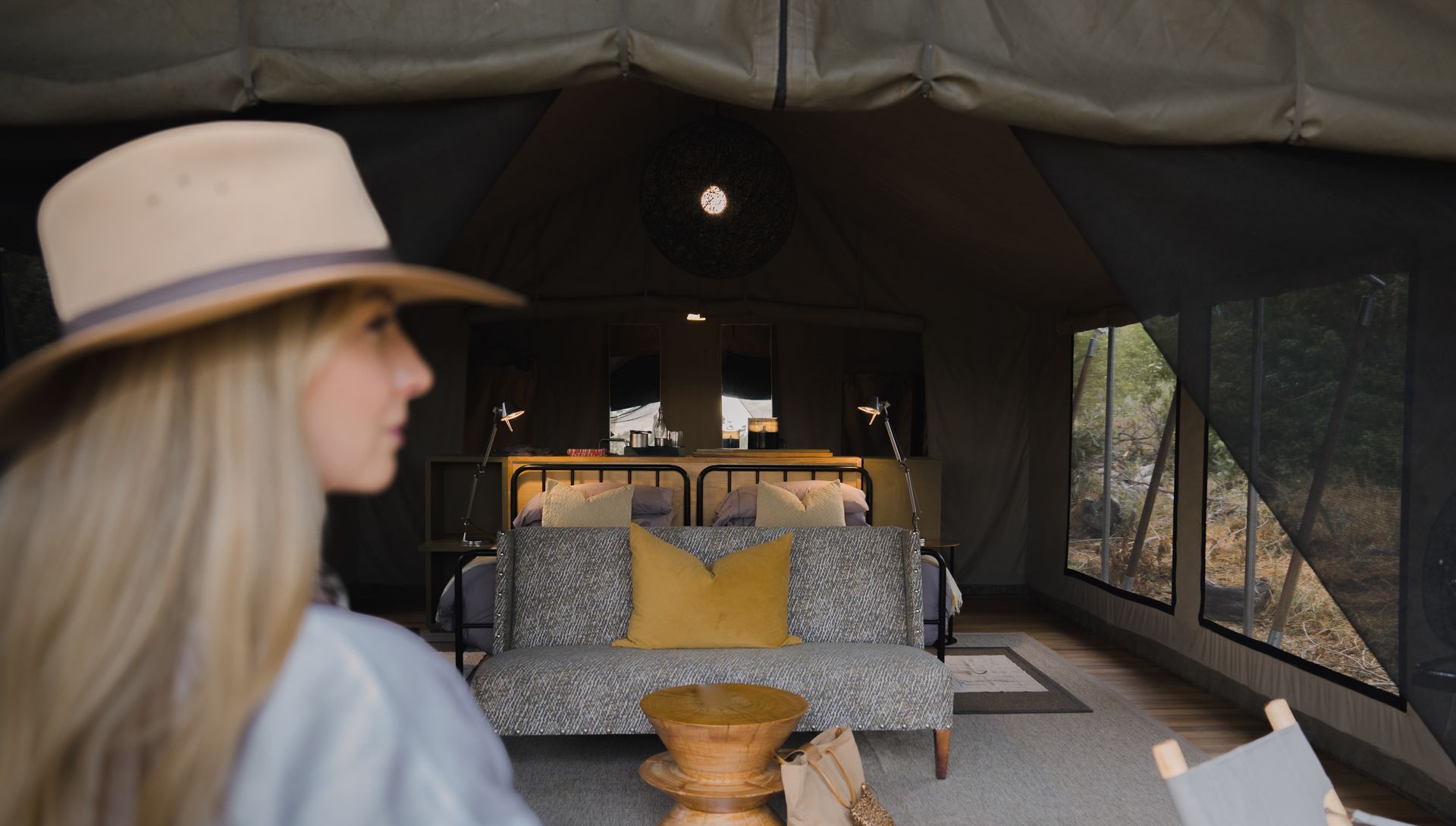 Woman in hat looks at safari tent interior with bed and seating.