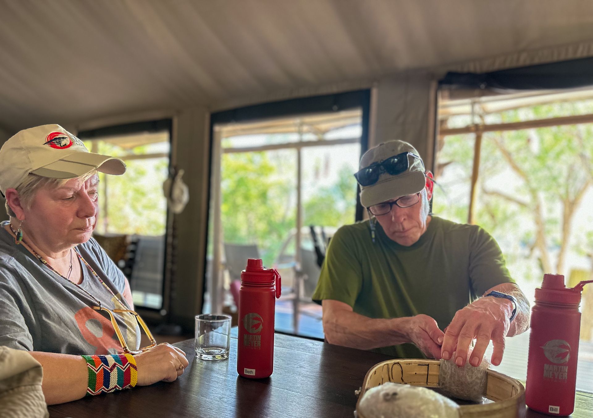 Two people seated at a table, one in a cap looking down, the other wearing a hat.