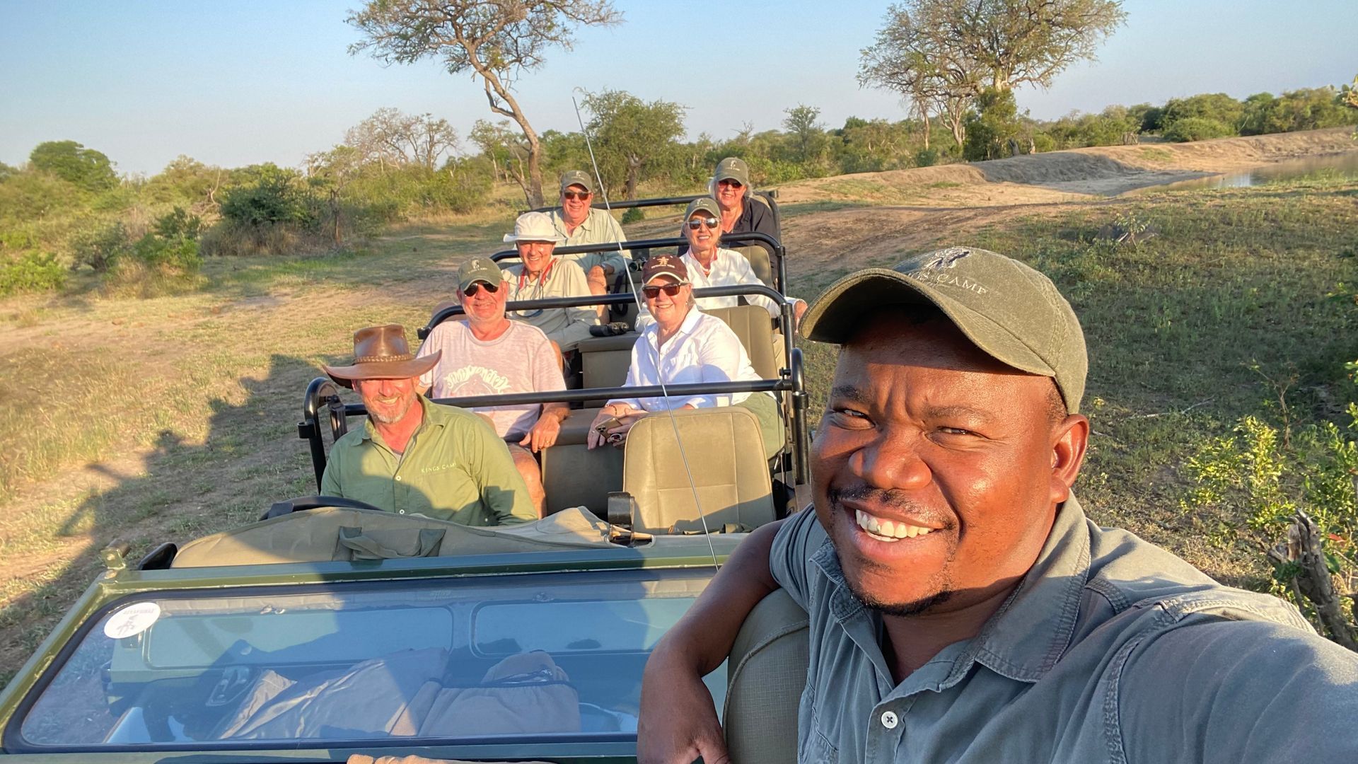Group riding in open safari vehicles on a dirt track, with one smiling driver in the foreground at sunset