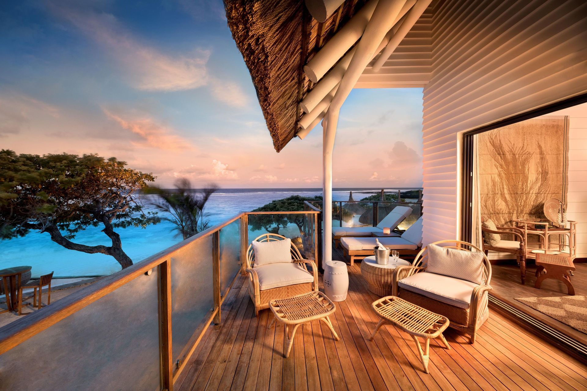 Balcony overlooking ocean, wooden deck, rattan furniture, sunset colors, open door to a room.