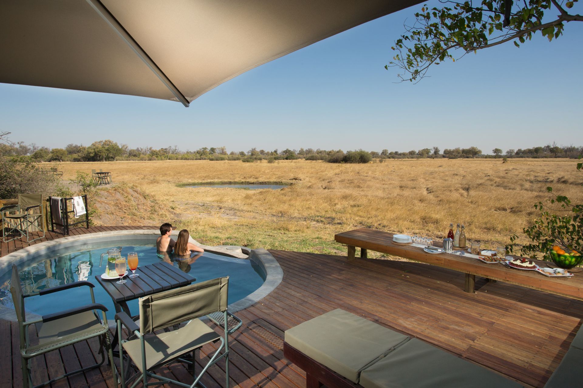 Couple relaxing in pool, outdoor setting with a table, drinks, and a view of a grassy field.