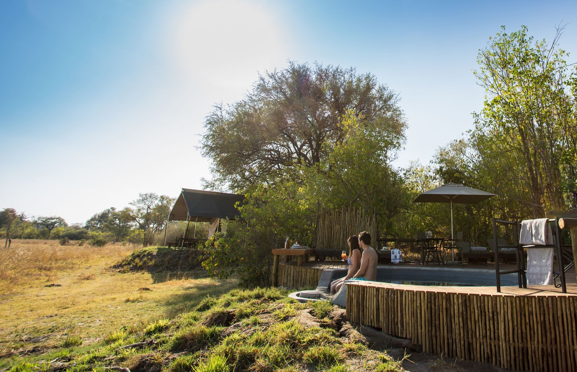People relaxing in a small pool next to a grassy field, trees, and a lodge on a sunny day.