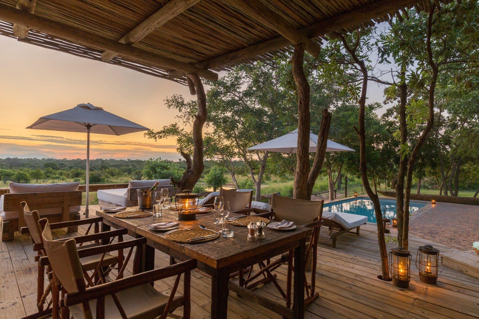 Outdoor dining area at sunset with a table set for guests, a pool, and a view of the African landscape.