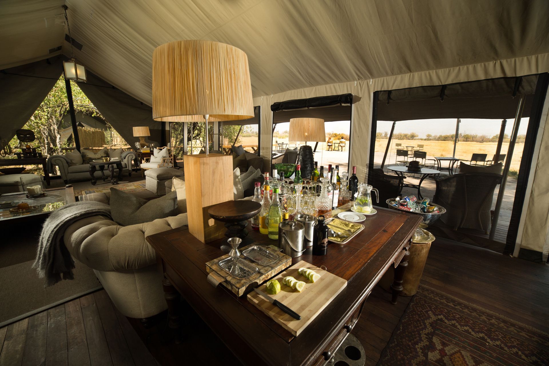 Lounge area inside a tent with a bar. A couch and table with drinks and snacks. Outdoor view through windows.