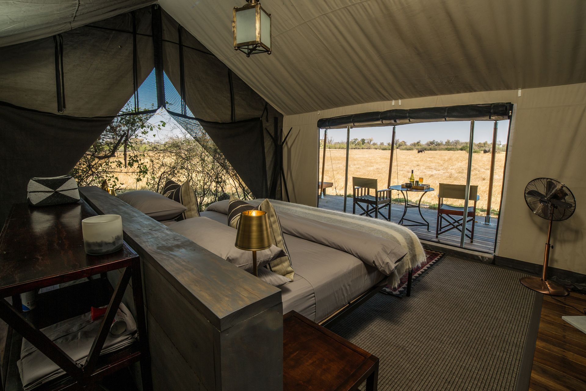 Safari tent interior with bed, table, and view of outdoor chairs on a sunny day.