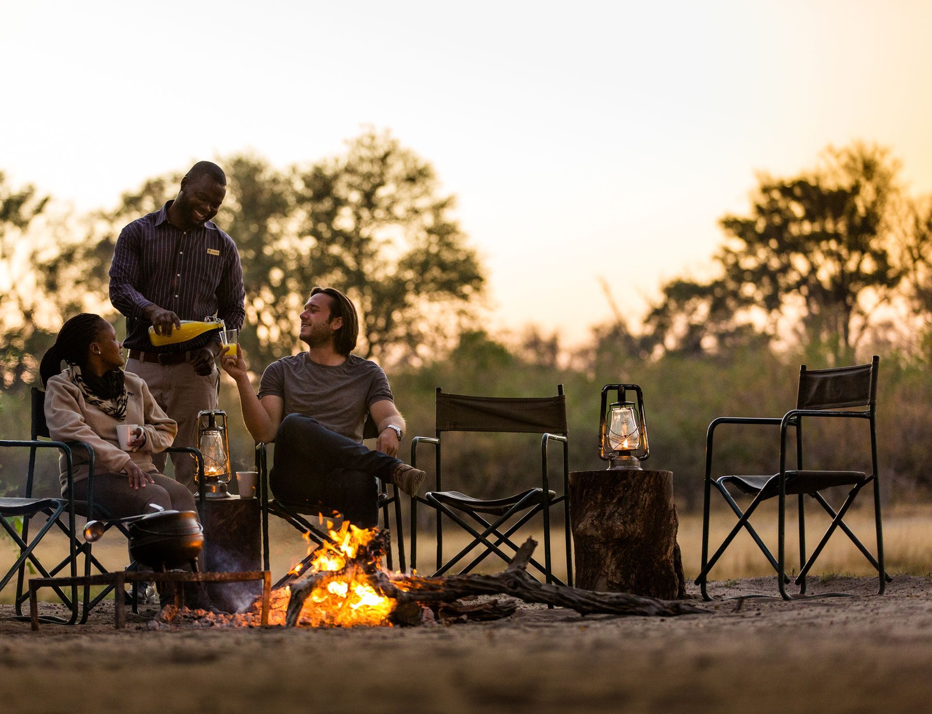 People around a campfire in an African safari setting, being served drinks by a staff member.