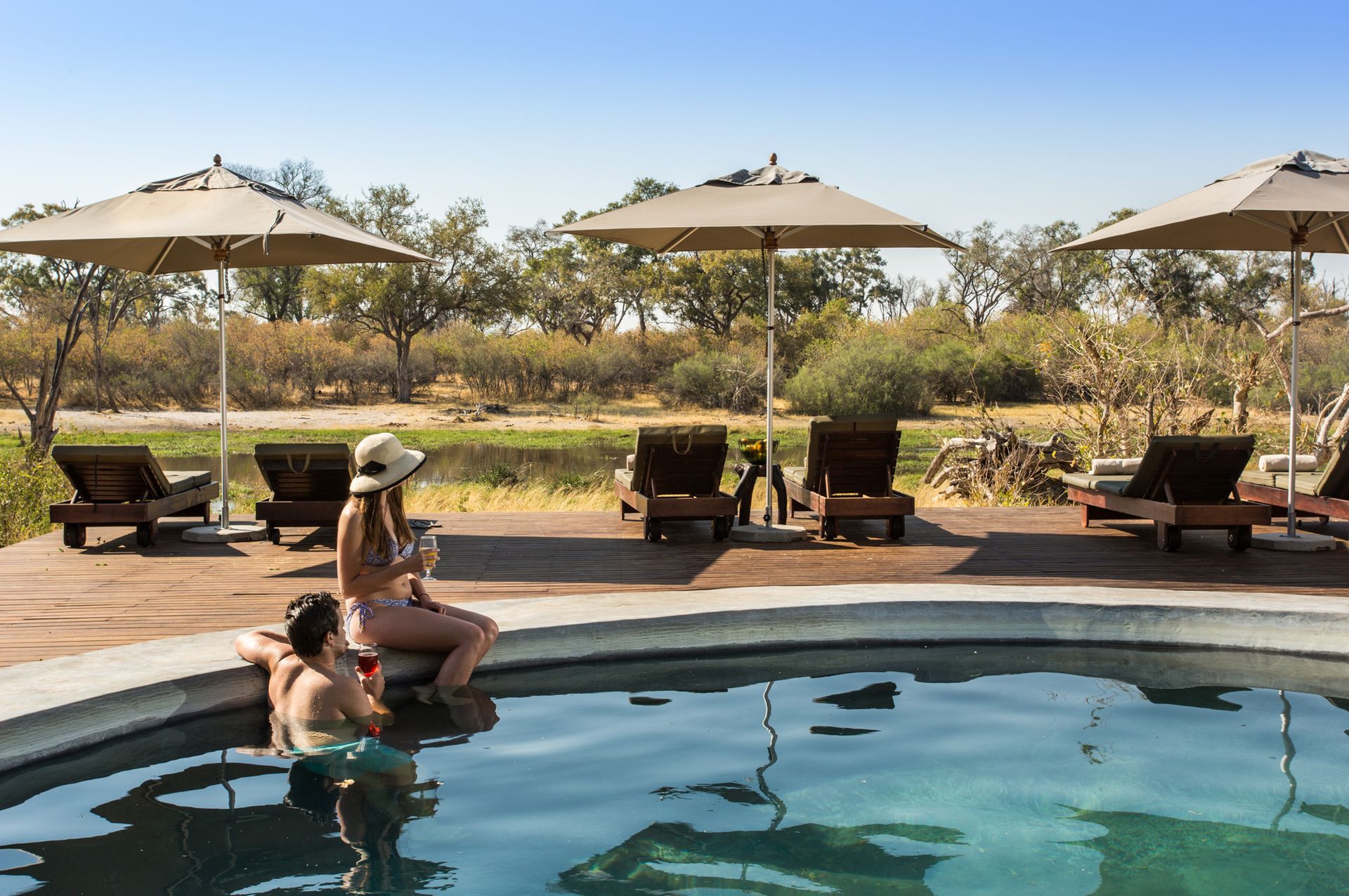 Couple relaxing in pool, woman wearing hat and holding drink, lounge chairs, umbrellas, outdoor setting.