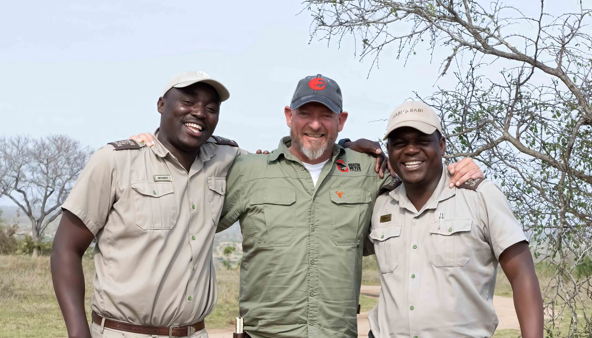 Three men smiling, posing in natural setting; one wears a red cap.