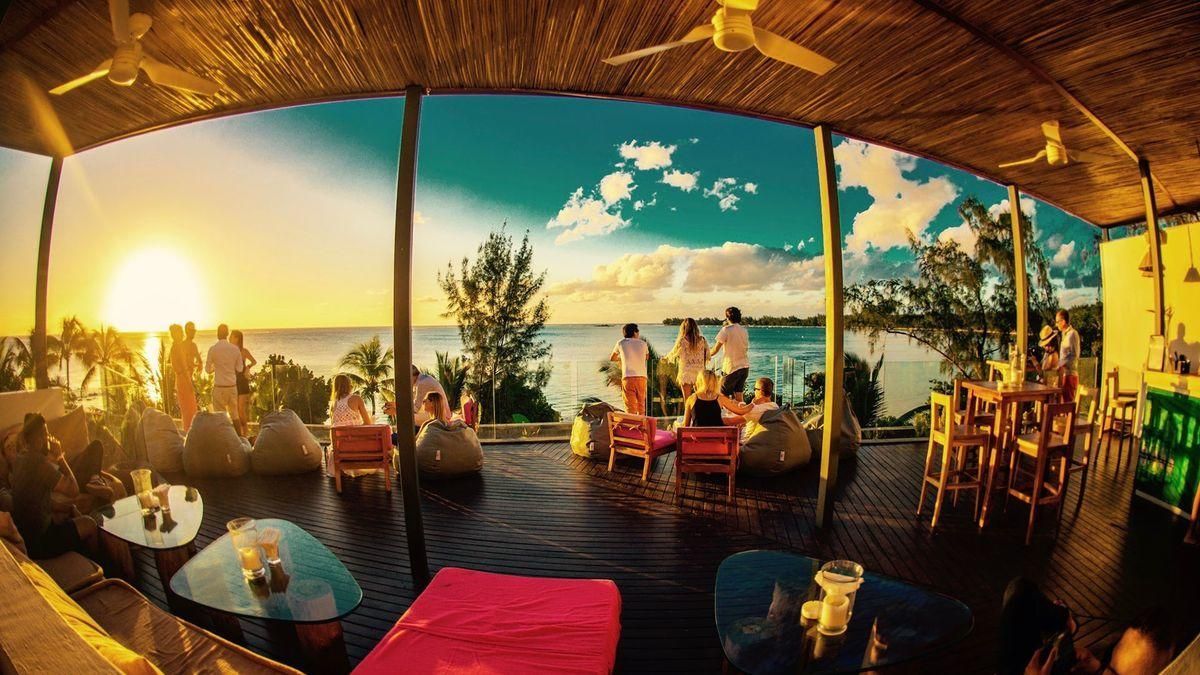 Sunset view from a beach bar with people, blue water, wooden deck, and colorful furniture.