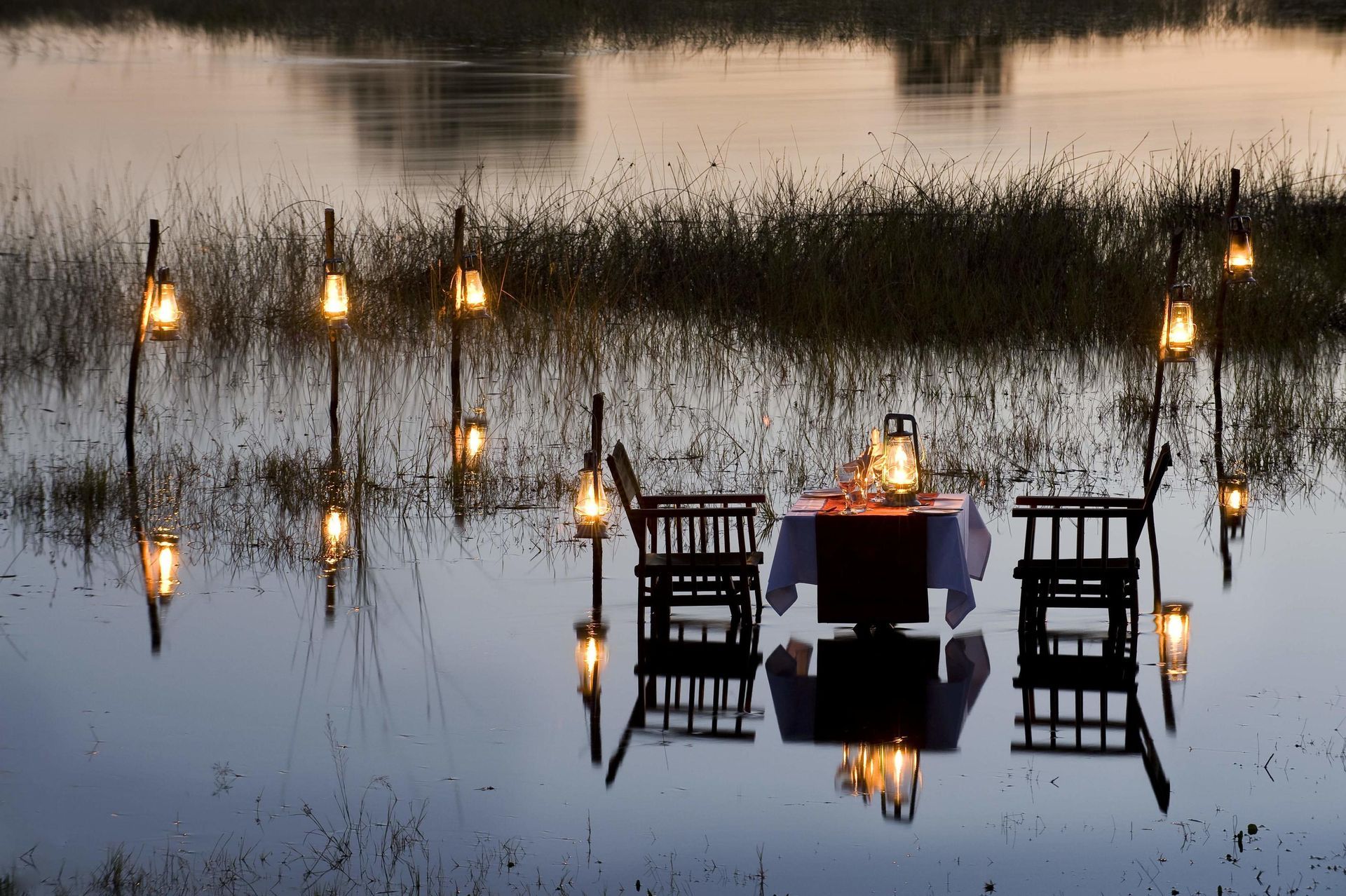 Romantic dinner setting in water, lanterns illuminating table and chairs.