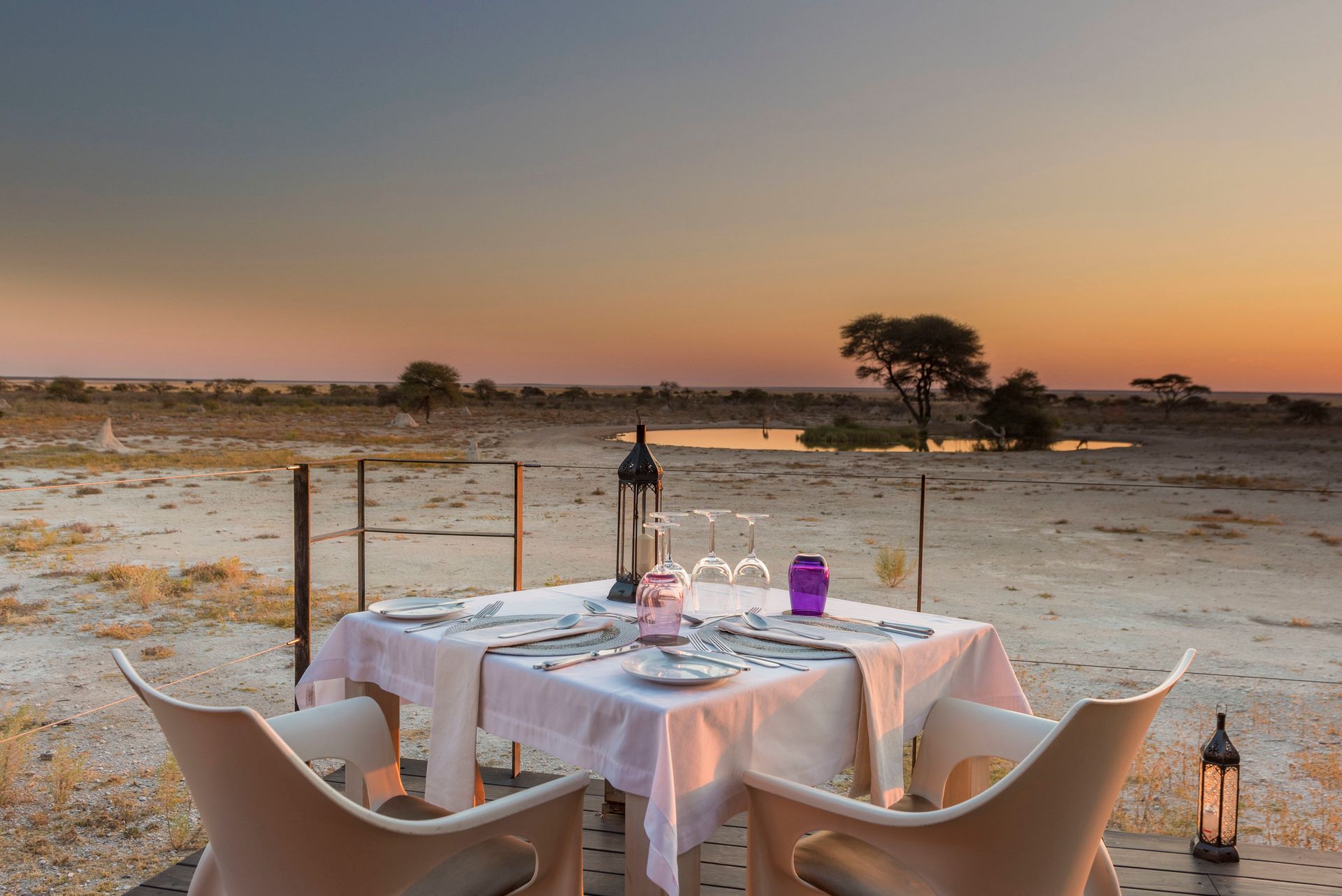 Sunset dining setup on a desert terrace with a table, chairs, lanterns, and white tablecloths