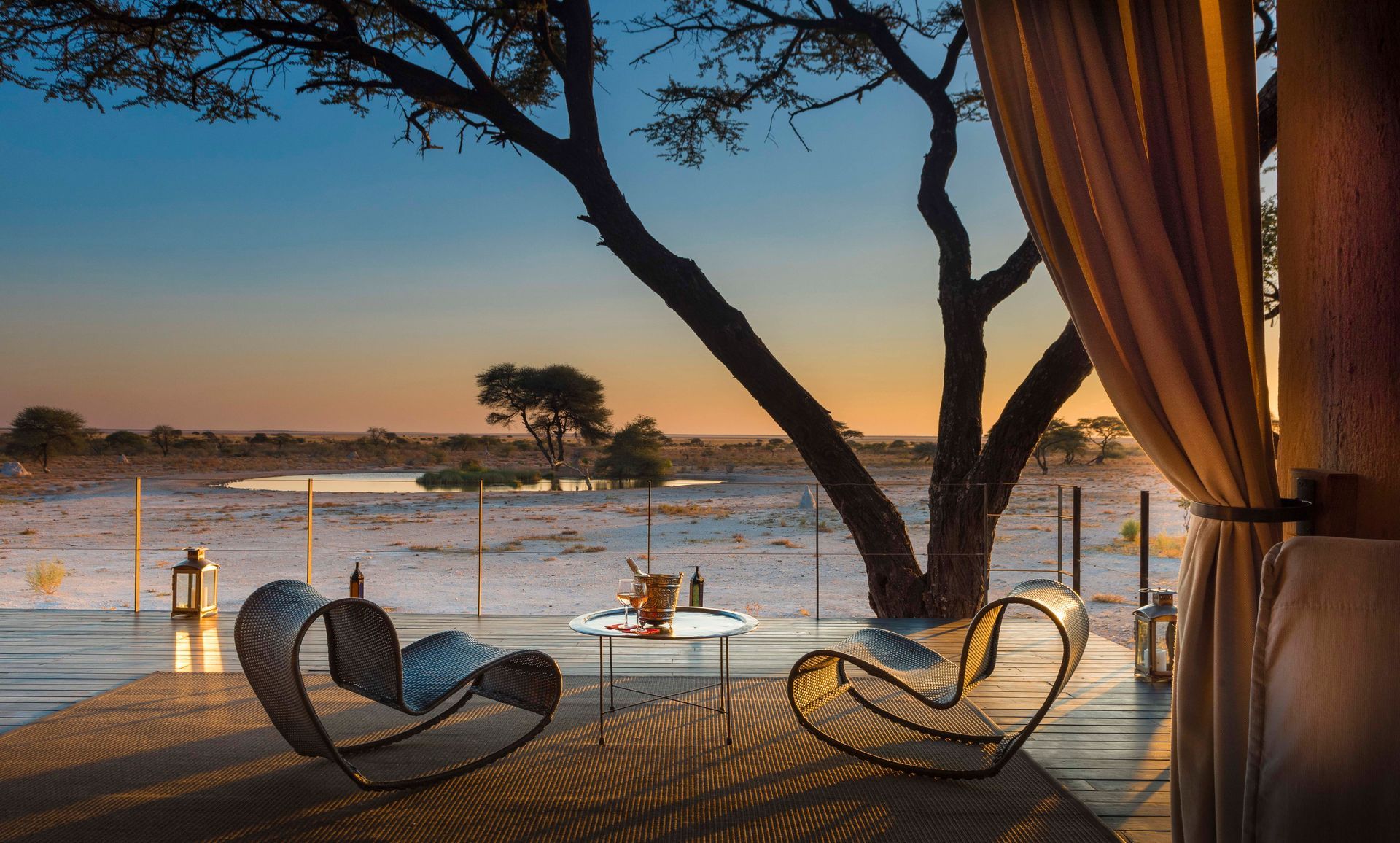Sunset over a lakeside deck with lounge chairs, lanterns, and a tree-framed view of the water