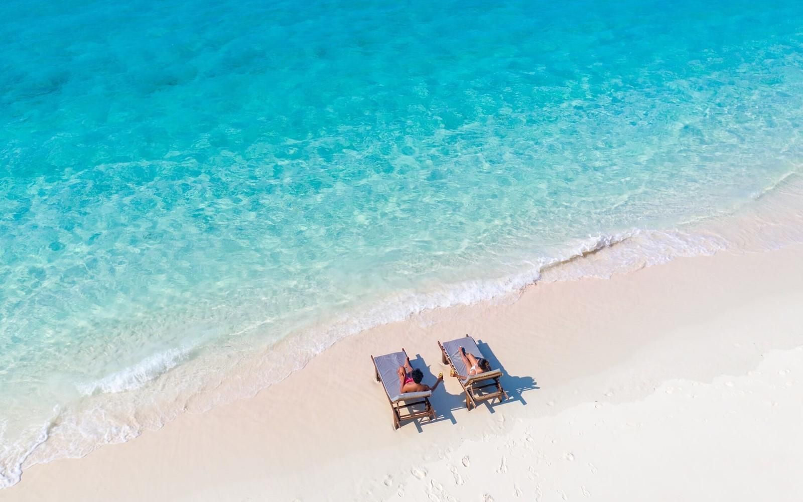 Two lounge chairs on a white sandy beach with turquoise water, an aerial view.