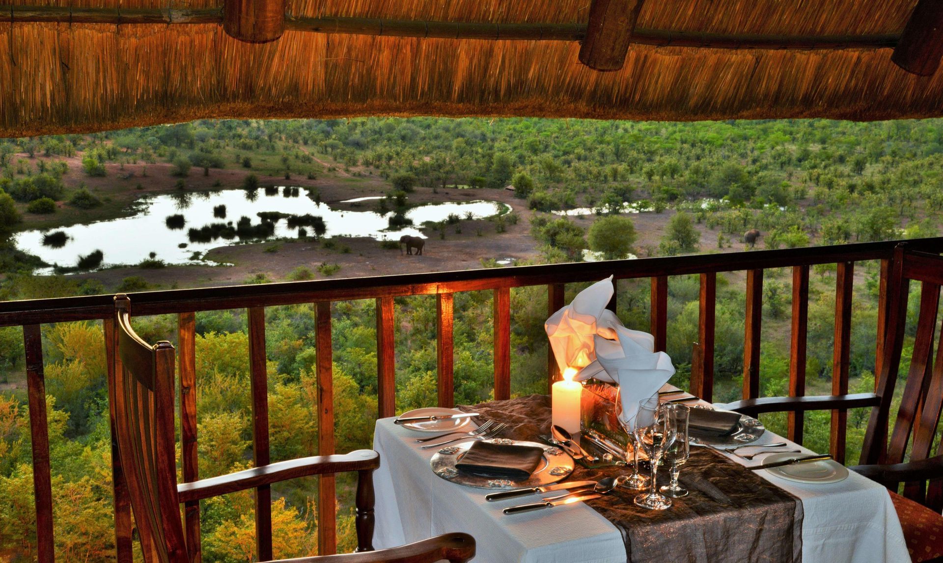 Romantic outdoor dining setting on a deck overlooking a river and forest, lit by candles at dusk.