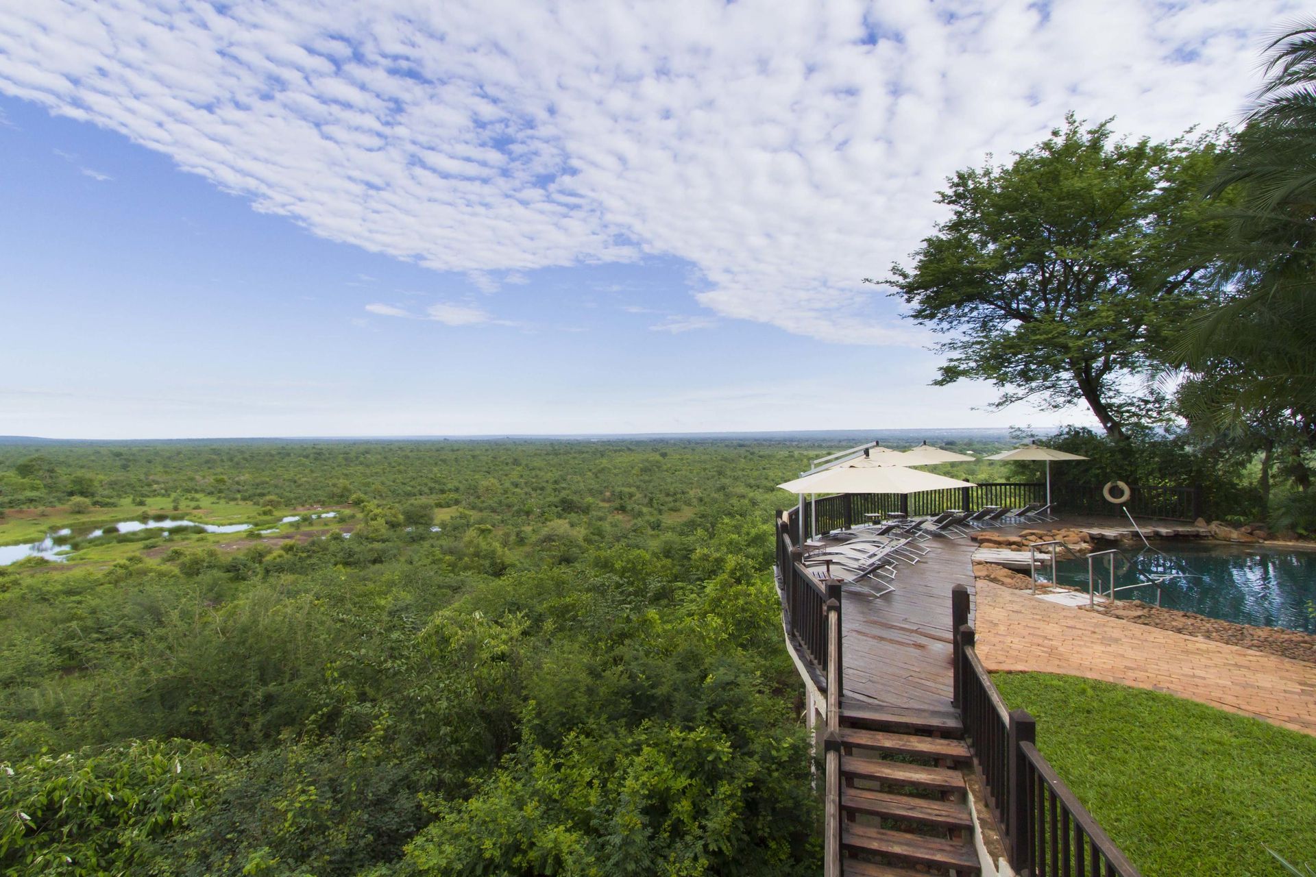 Terrace overlooking a vast green landscape, trees, and cloudy blue sky. A pool and some tables are visible.