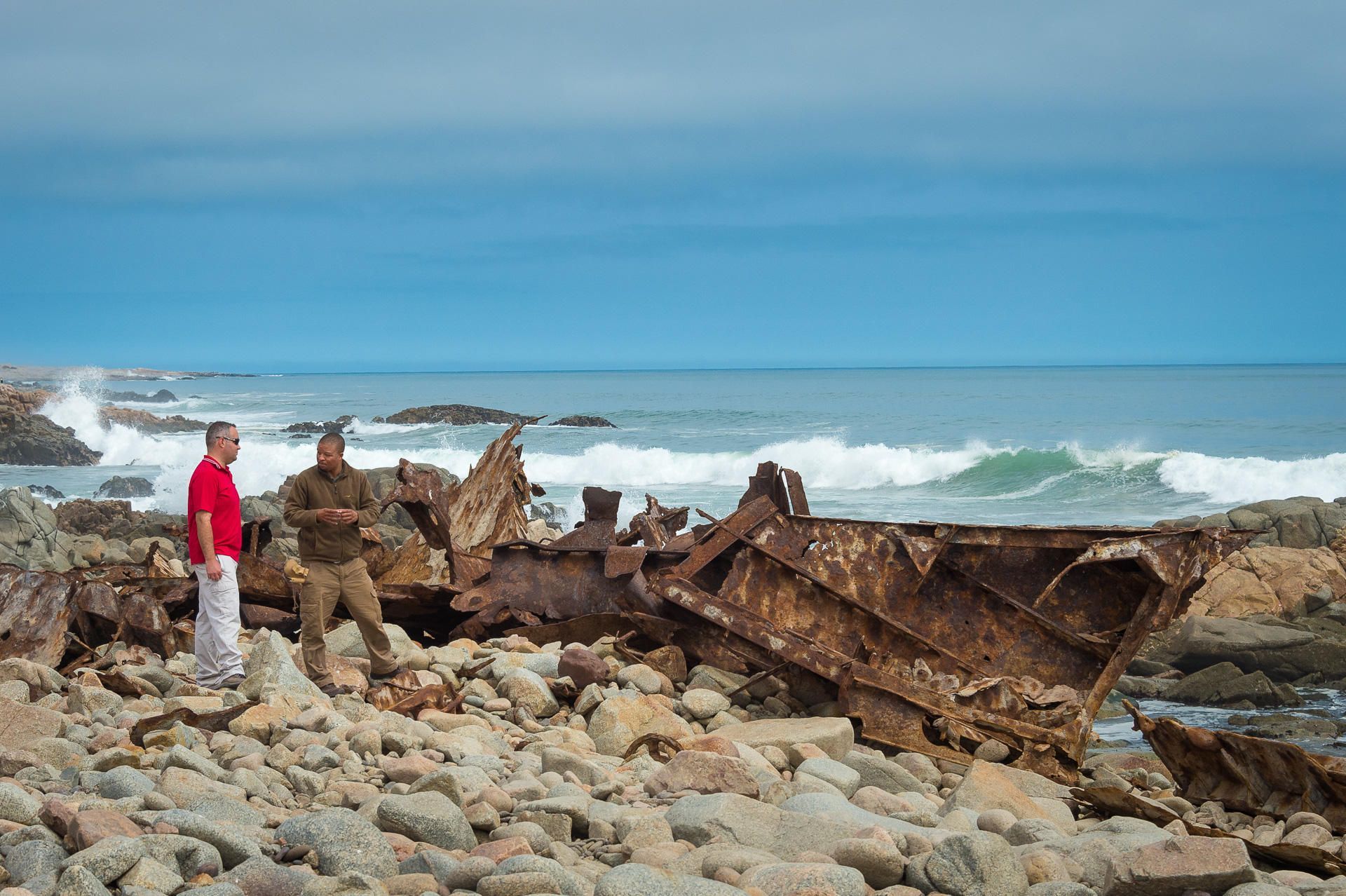 Two men are standing on a rocky beach next to a boat wreck. Namibia Safari