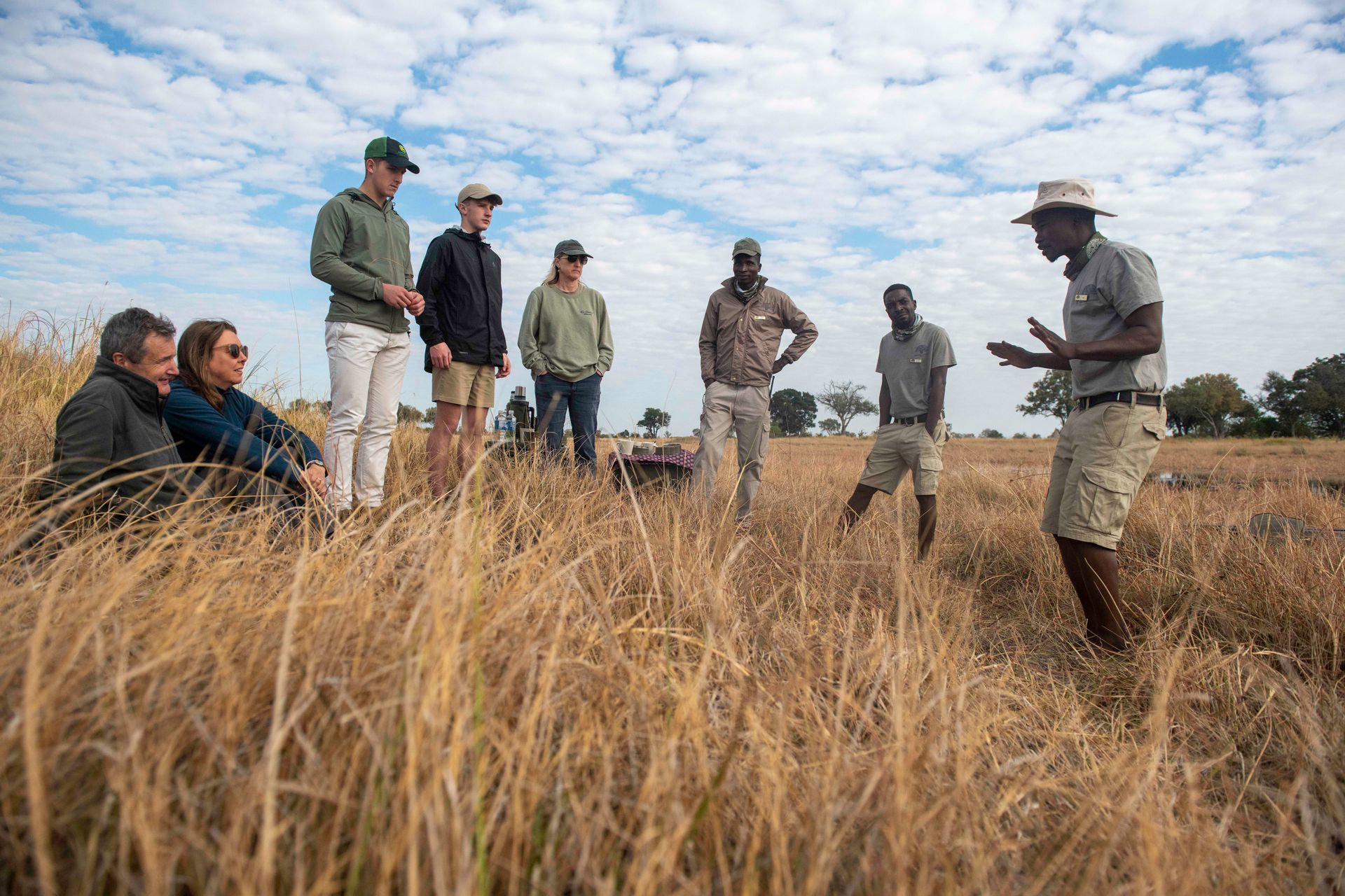 Group of people on a field trip, listening to a guide. Tall, dry grass and blue sky.