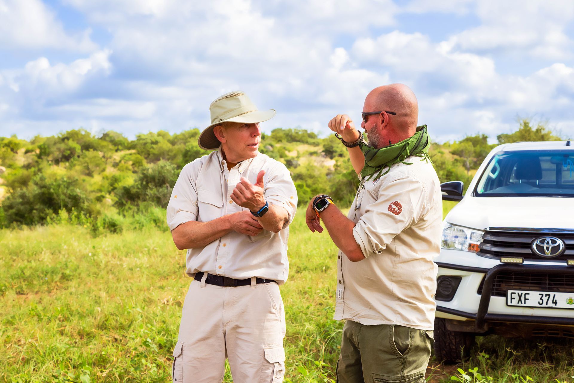 Two men in safari attire conversing outdoors near a white SUV and greenery. One gestures while the other listens.