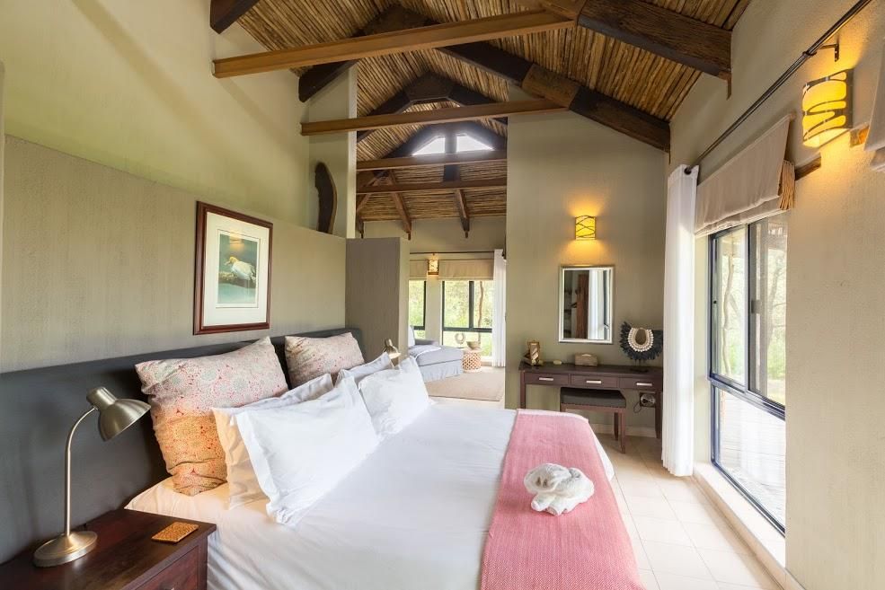 Bedroom with king bed, pink blanket, wooden ceiling, and large window overlooking trees.