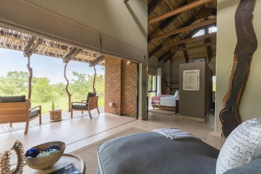 Luxury lodge interior with outdoor deck overlooking a green landscape. Exposed beams, brick wall. Grey couch and chairs.