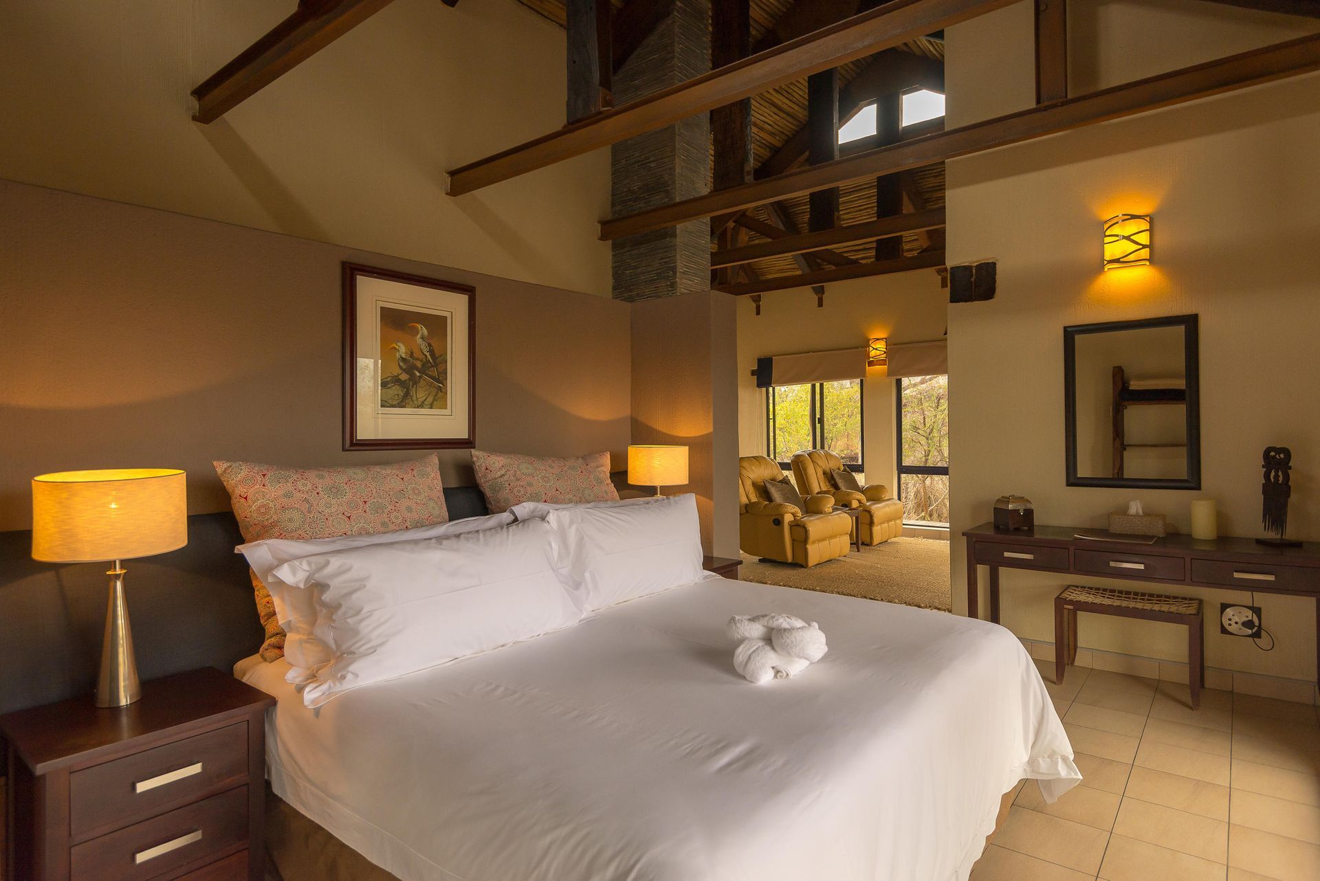Bedroom with a large bed, nightstands, and a desk, open to a living area with armchairs.