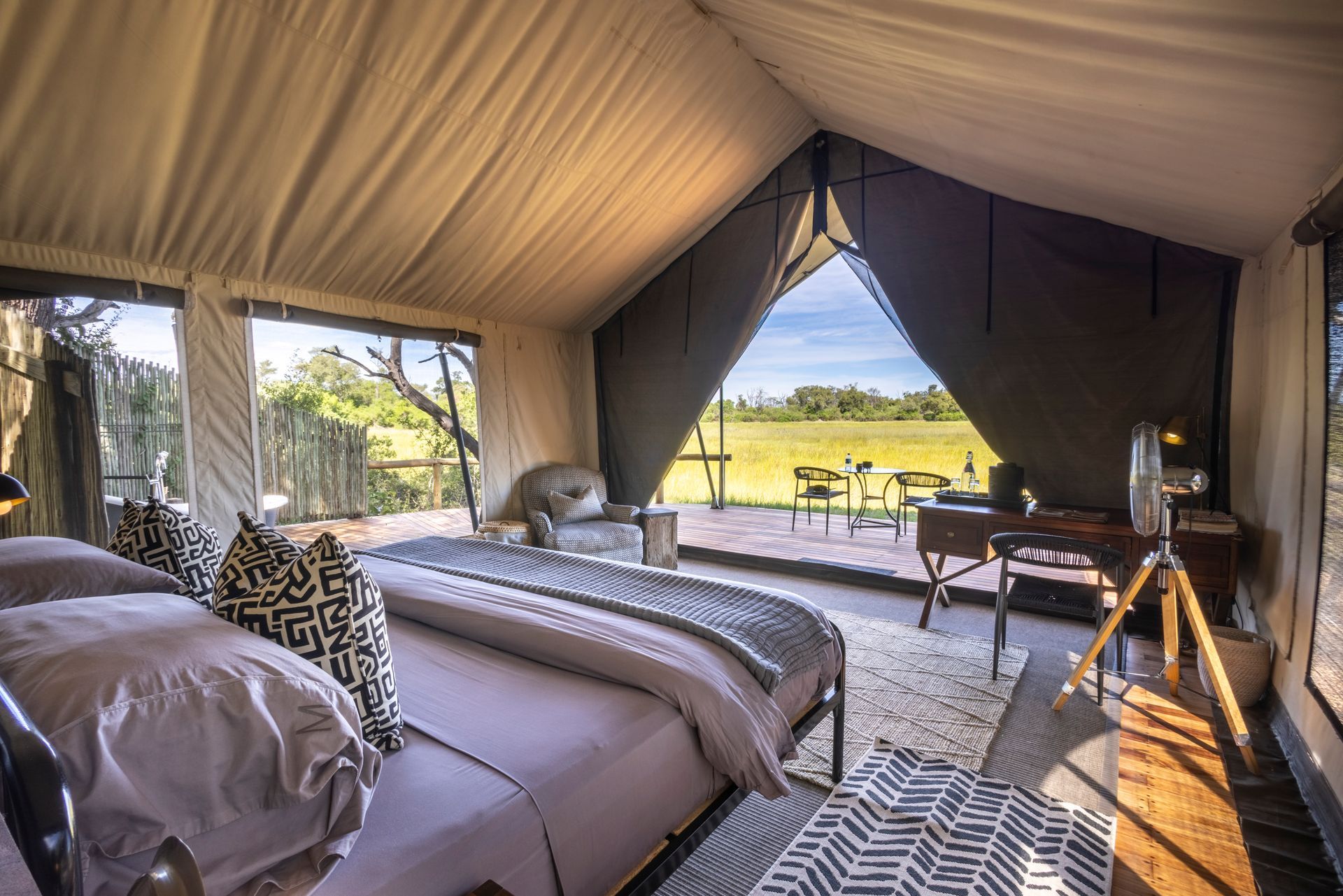 Interior of a glamping tent with a bed, desk, and open doorway leading to outdoor seating overlooking a scenic view.