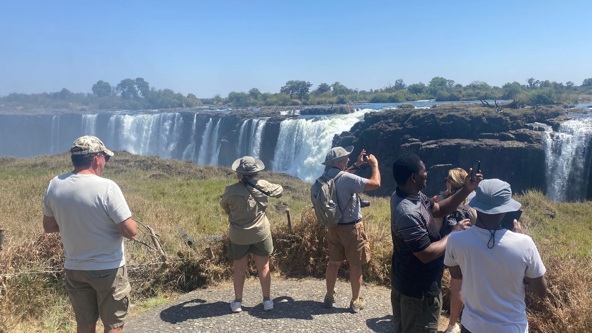 Tourists viewing a wide waterfall from a grassy lookout on a sunny day