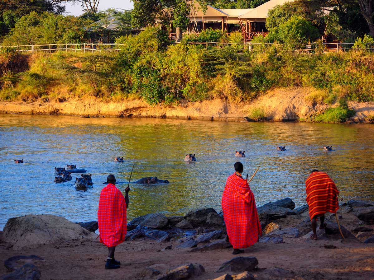 Three Maasai men in red robes watch hippos in a river near a lodge.