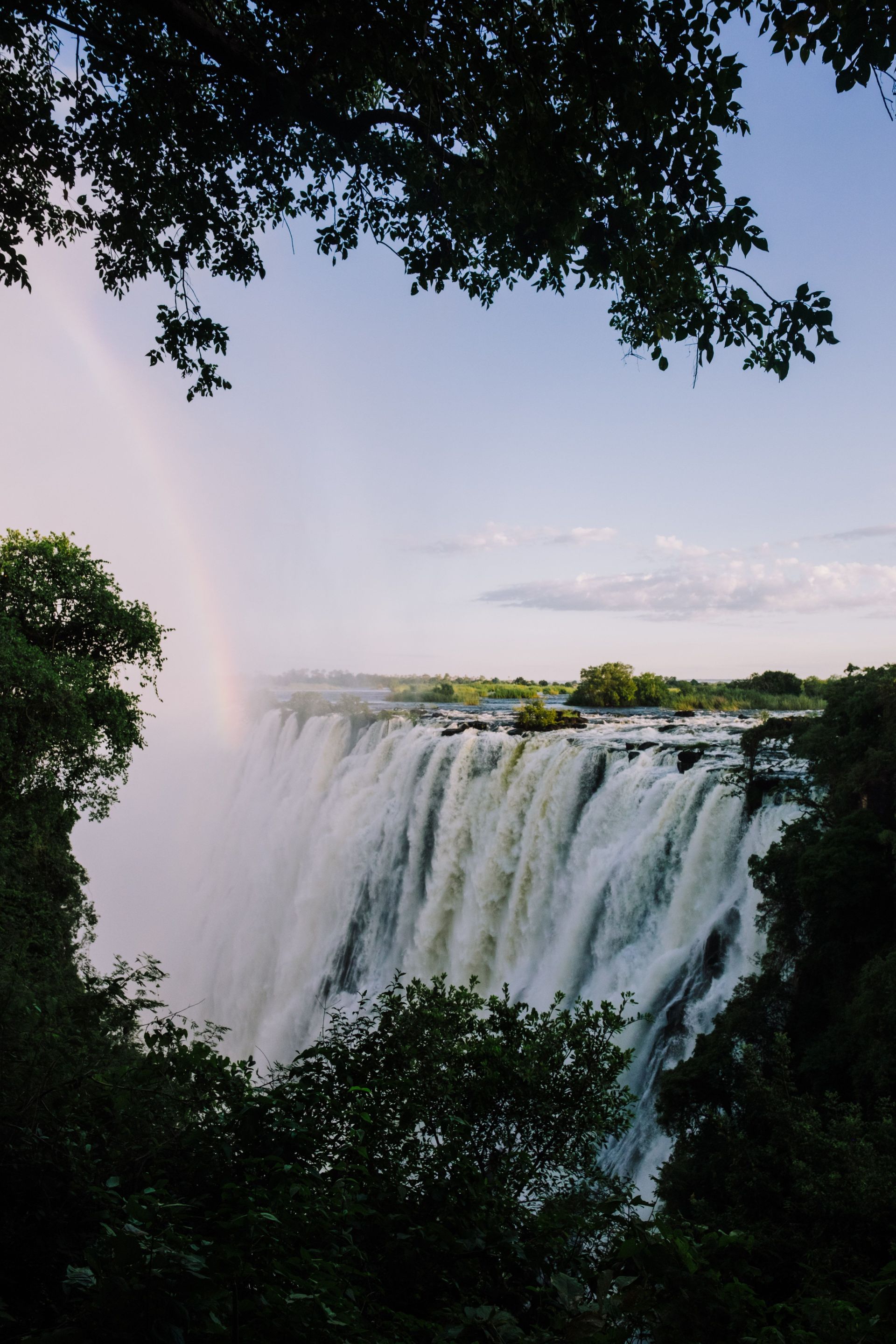 Wide waterfall cascading over a cliff, framed by dense green trees under a pale sky.