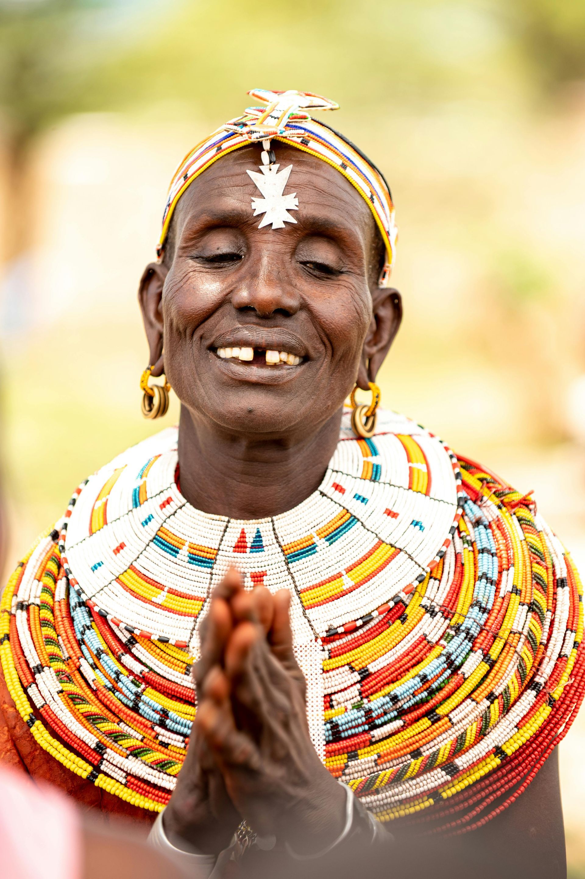 Person with a beaded collar and headdress, hands clasped, smiling with eyes closed.