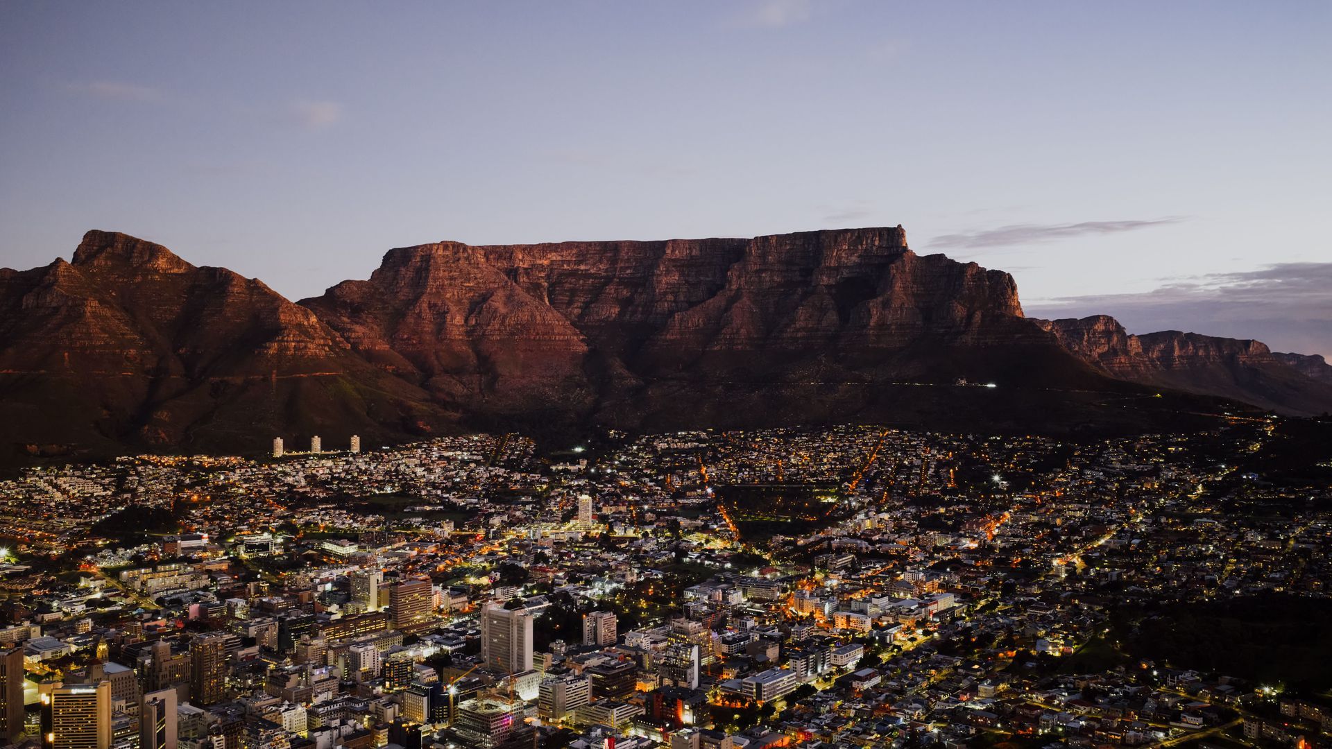 City lights below a red sandstone mountain range at dusk, with Table Mountain in the background