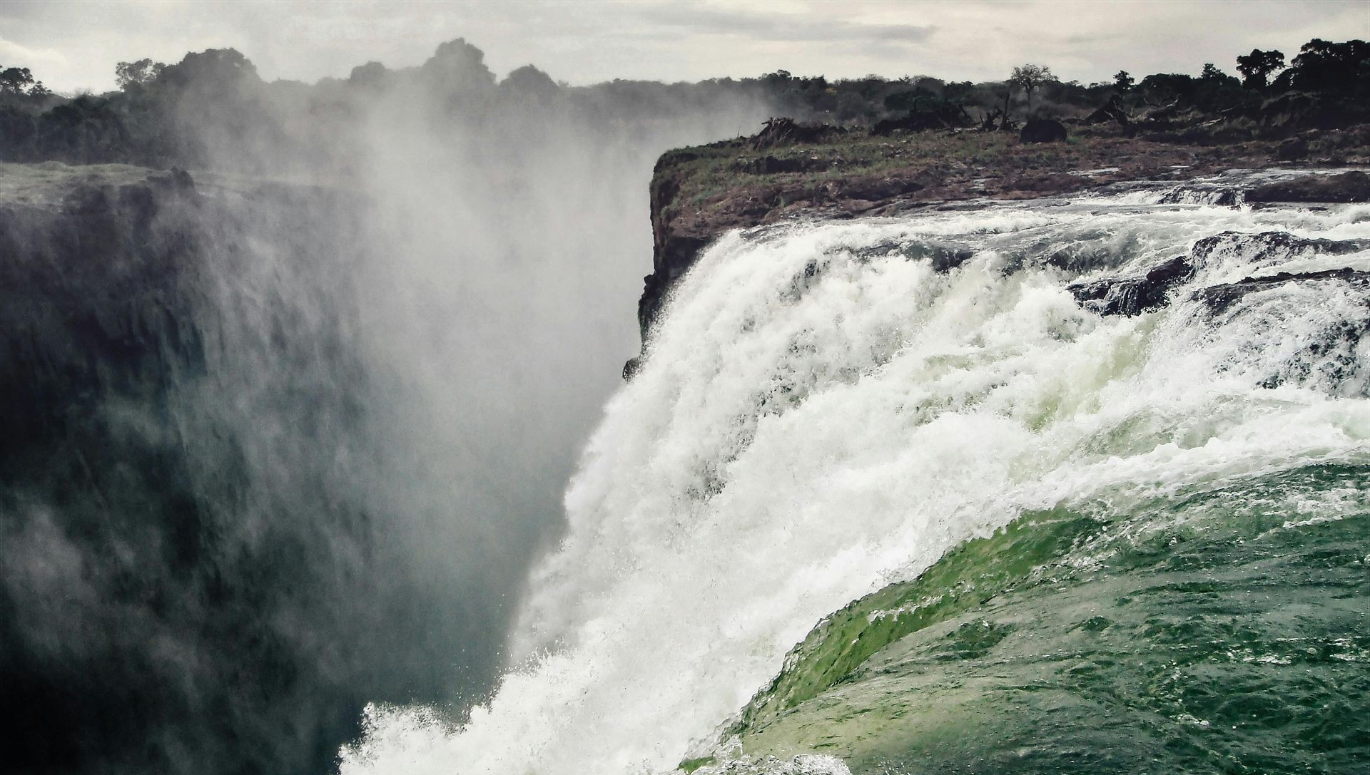 Waterfall cascading over a cliff, creating mist; green water and vegetation visible.