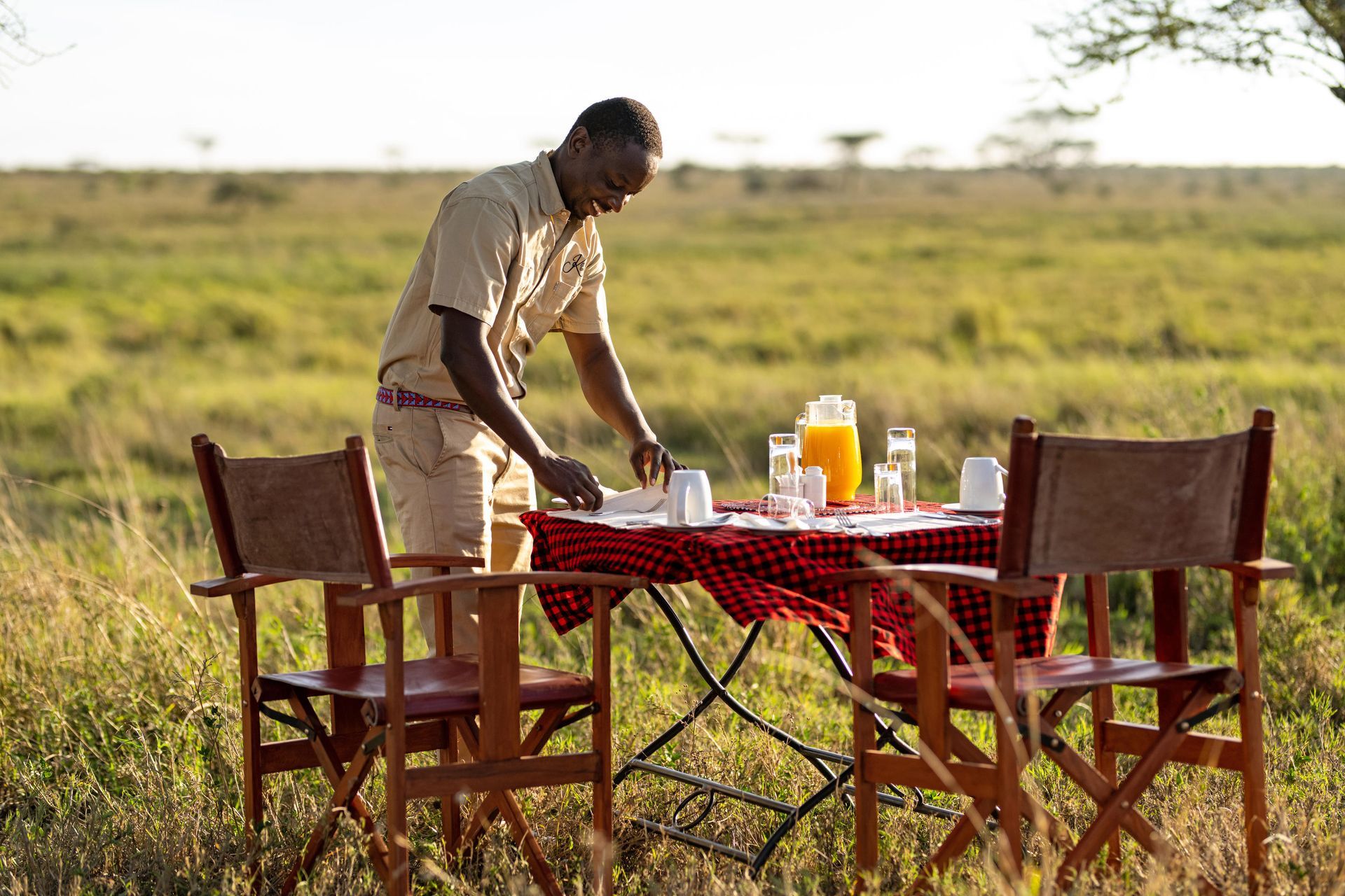 Person setting a table with drinks in a grassy outdoor savanna setting