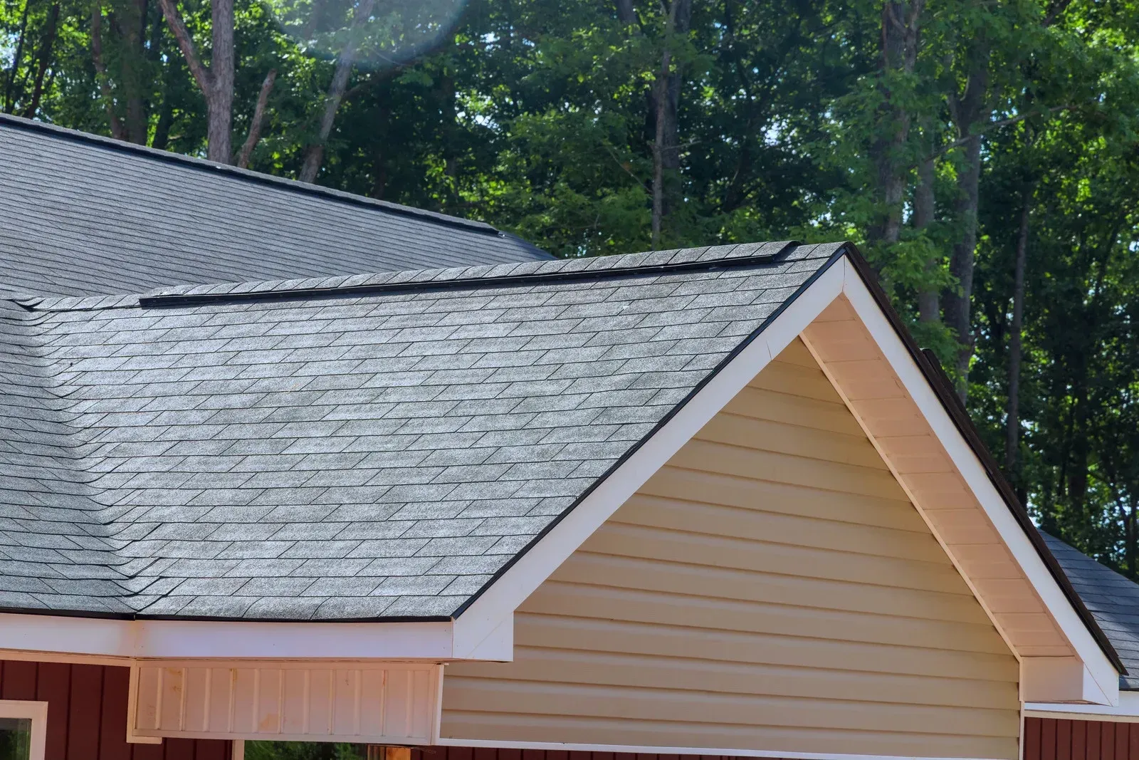 Gray shingled roof on a tan house with white trim, backed by tall green trees
