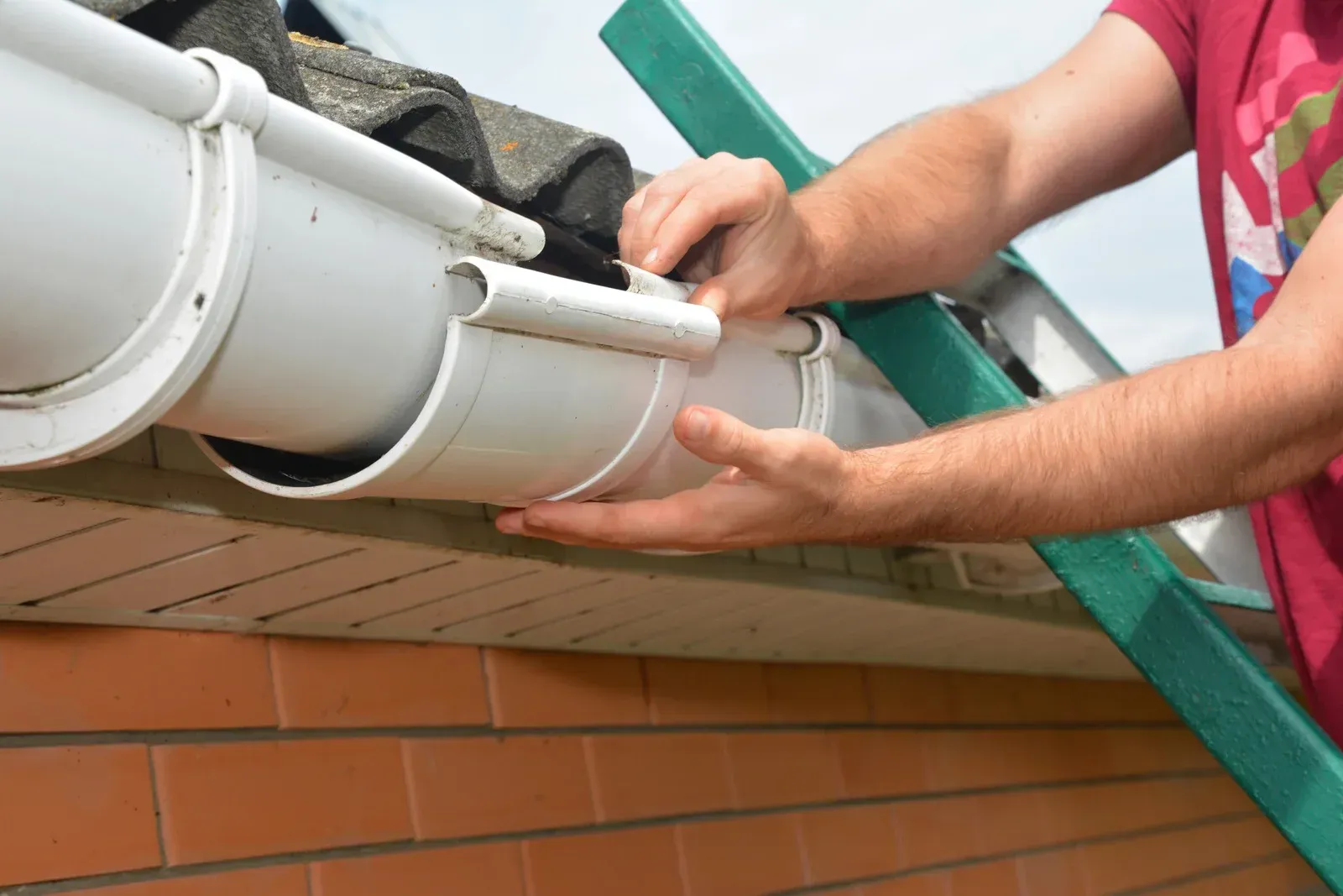 Hands installing white gutter on a brick house roof, with ladder nearby.
