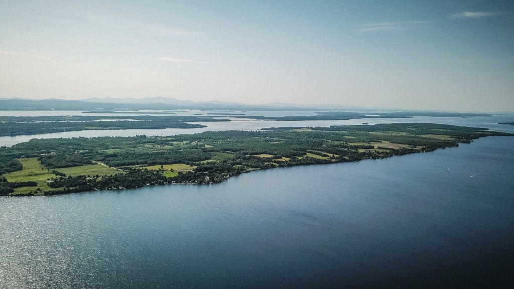 Aerial view of a long peninsula with farmland and forest extending into a wide lake, with distant mountains