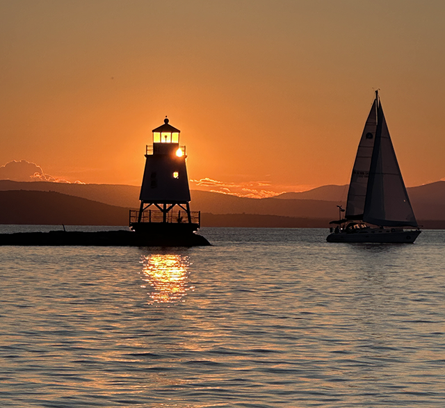Lighthouse silhouetted against an orange sunset, with a sailboat passing on the lake