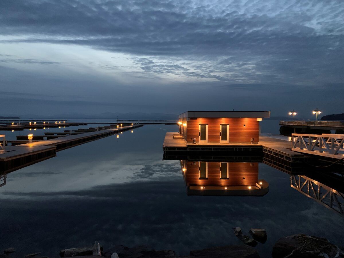Wood-clad floating building lit up at dusk on still water, with long docks and a cloudy sky