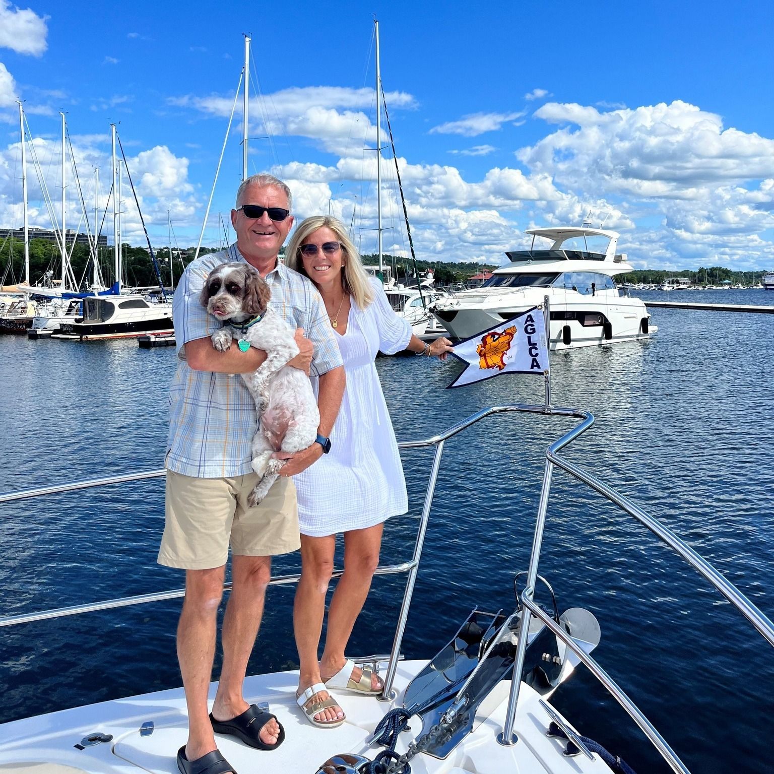 Couple holding a small dog on the bow of a boat, with sailboats and a yacht behind them on a sunny day