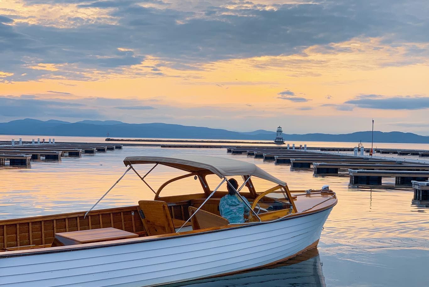 Classic wooden boat with a canopy on calm water at sunset, with floating docks, a lighthouse, and mountains behind